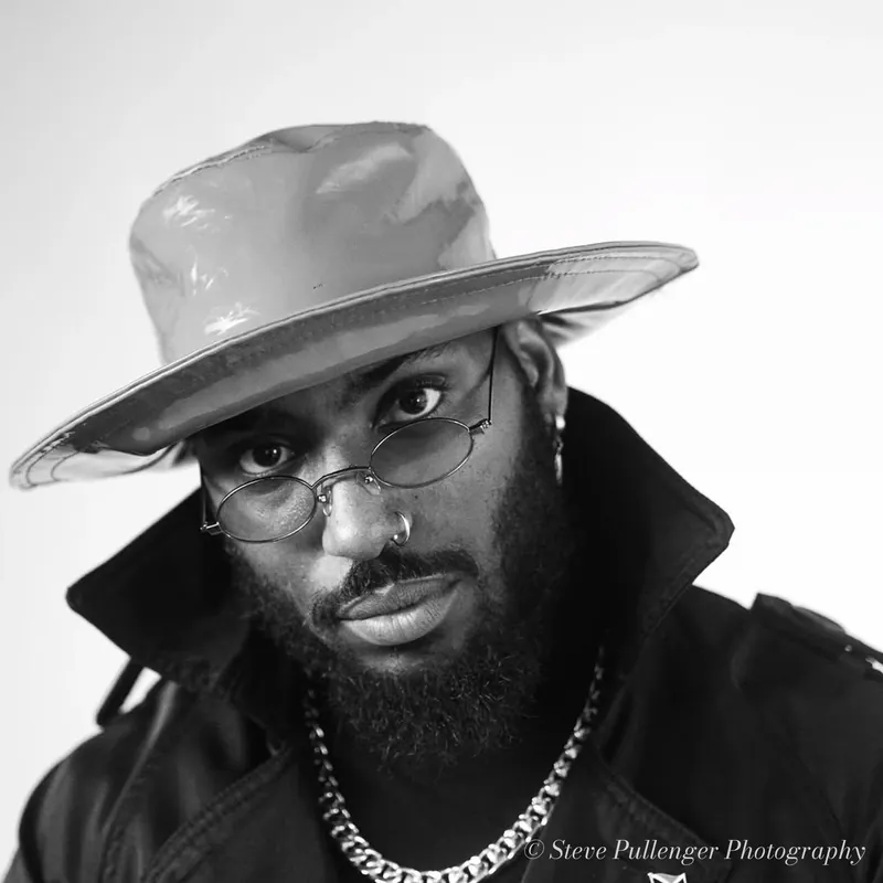 Black and white studio portrait of a bearded man in a patent wide-brim hat and trench coat wearing a chunky chain